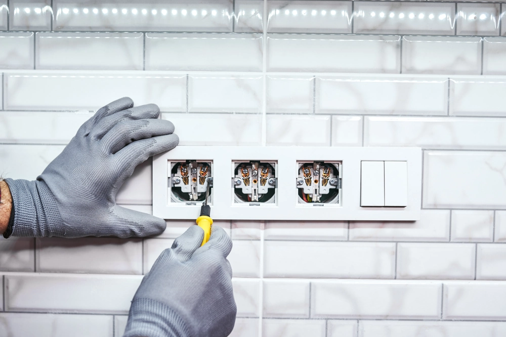 Montaż gniazdek: elektryk w rękawicach dokręca trójgniazdo elektryczne na ścianie wykończonej białymi płytkami