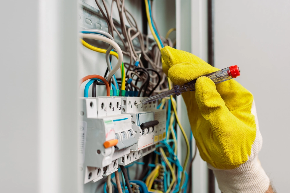 Ręka w żółtej rękawicy sprawdza napięcie w rozdzielnicy elektrycznej – pomiary instalacji elektrycznych